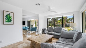 A spacious living room with a large grey sofa, a wooden coffee table, and a ceiling fan. There is a framed plant on the wall and a view of palm trees outside. - Yacht Harbor Oasis: Beach Bliss with Private Pool  (West Palm Beach)