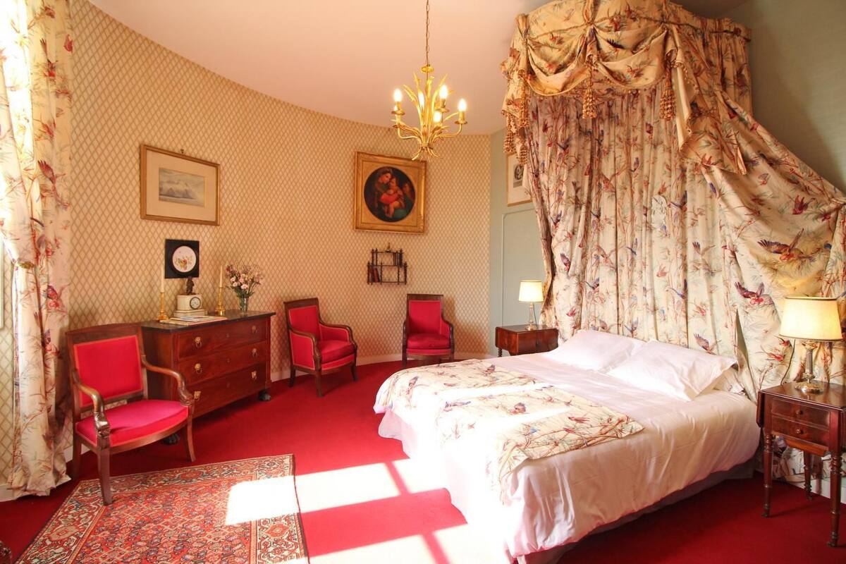 Ein Schlafzimmer mit Himmelbett, roten Stühlen, einer Holzkommode und einem gemusterten Teppich.