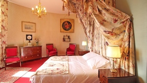 Un dormitorio con una cama grande, sillas rojas, un aparador, una araña y una cortina con motivos florales.