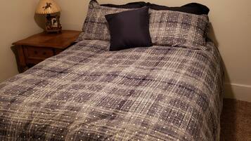 A bedroom with a patterned bedspread, a dark pillow, a nightstand with a lamp, and a window with blinds.