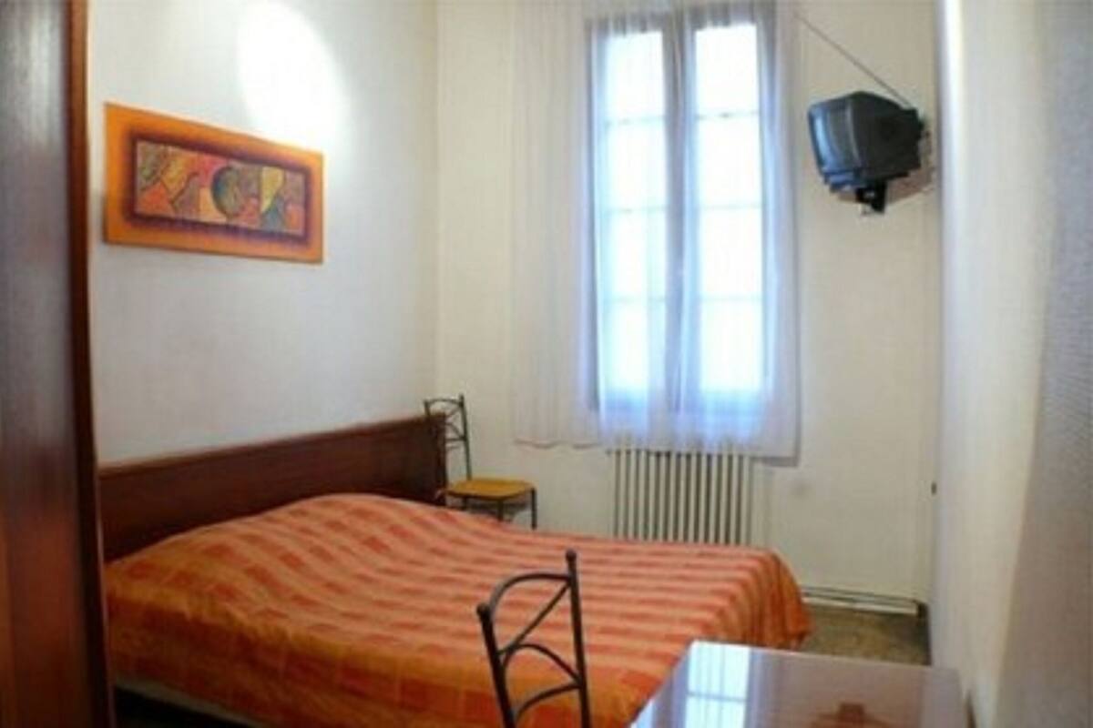 A small, simple room with a bed, a chair, a table, and a window with curtains.