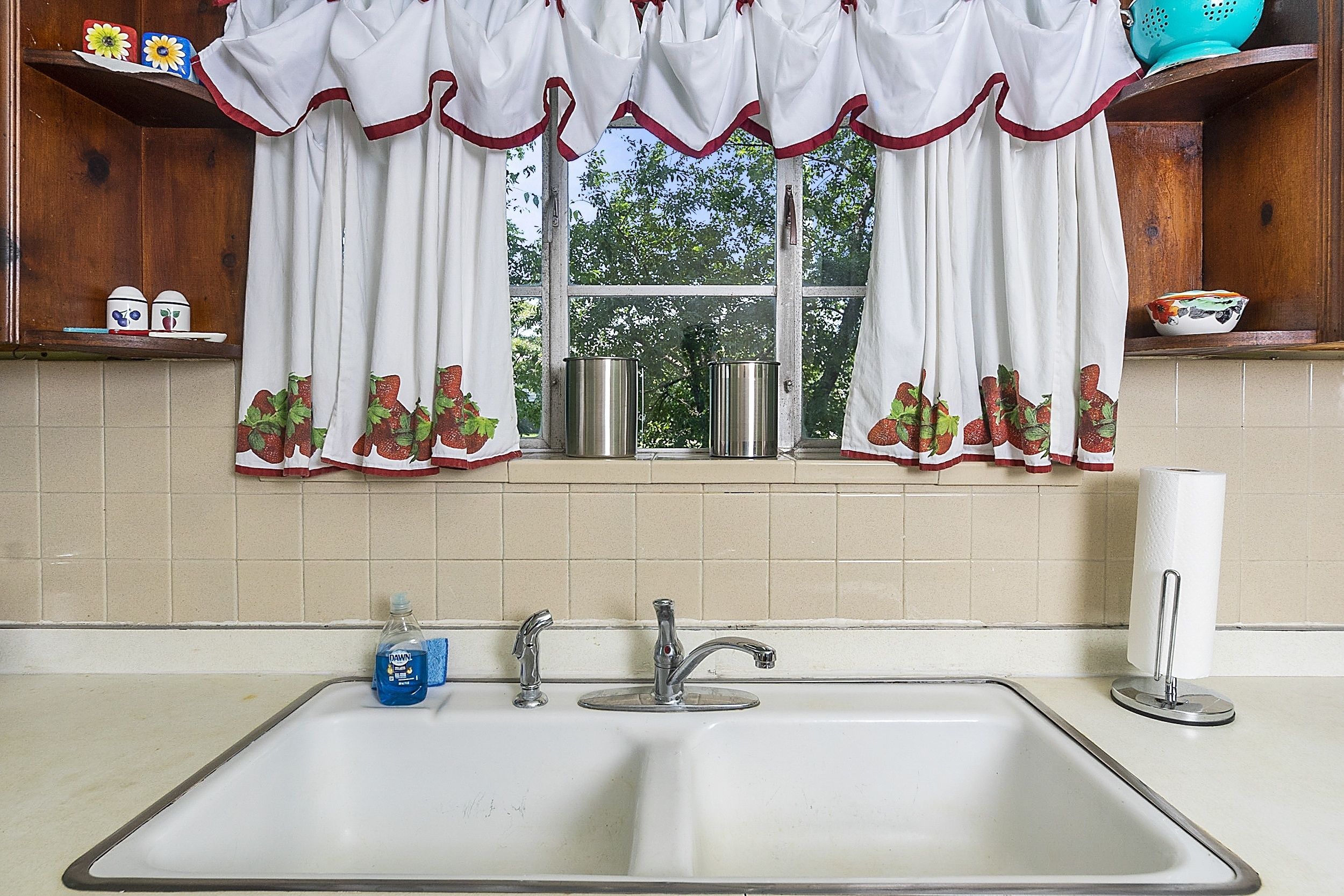 A kitchen with a double sink, a window with curtains, and a shelf with kitchen items.