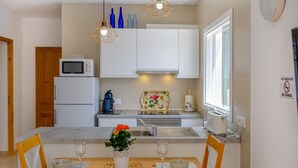 A modern kitchen with white cabinets, a microwave, a kettle, and a sink. There are two hanging pendant lights, a window with a view, and a dining area with a table and chairs. - Castell des Paradís - Family villa near the sea (Portopetro)