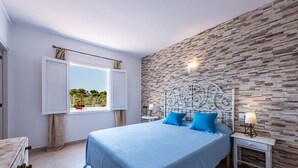 A bedroom with a stone-patterned wall, a bed with blue bedding, a nightstand with a lamp, and a window with curtains. - Castell des Paradís - Family villa near the sea (Portopetro)