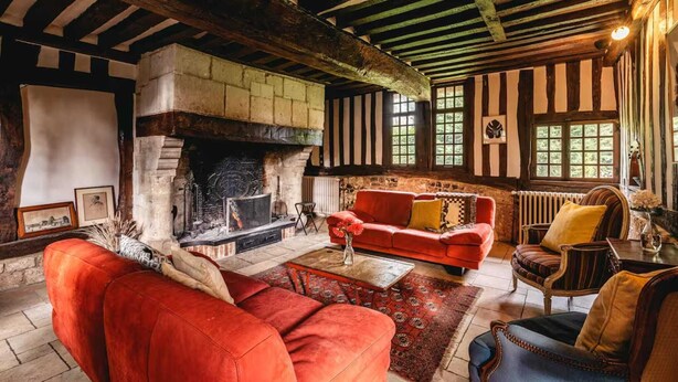 Una habitación rústica con vigas de madera a la vista, una chimenea de piedra, un sofá rojo y una alfombra estampada.
