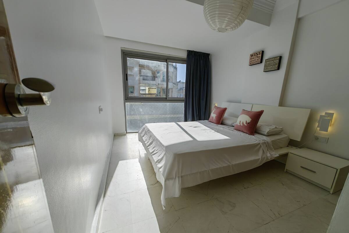 A modern bedroom with a large bed, a nightstand, a bedside table, a window with curtains, and a view of buildings outside.