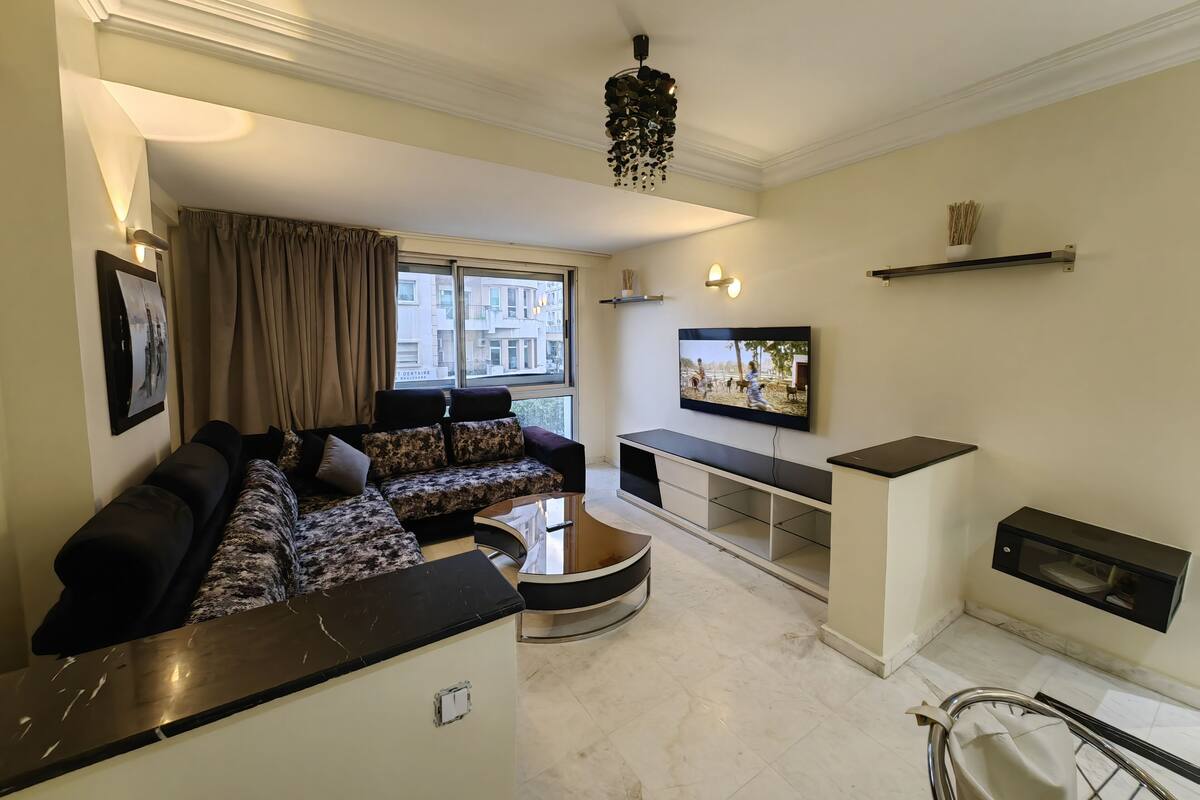 A modern living room with a sectional sofa, a TV mounted on the wall, and a dining area with a built-in oven.