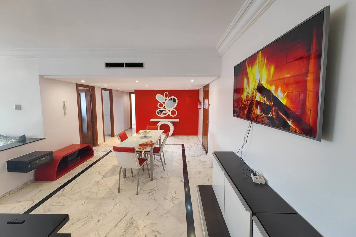 A modern dining area with a white table and red chairs, a red accent wall, and a large painting of a fire.