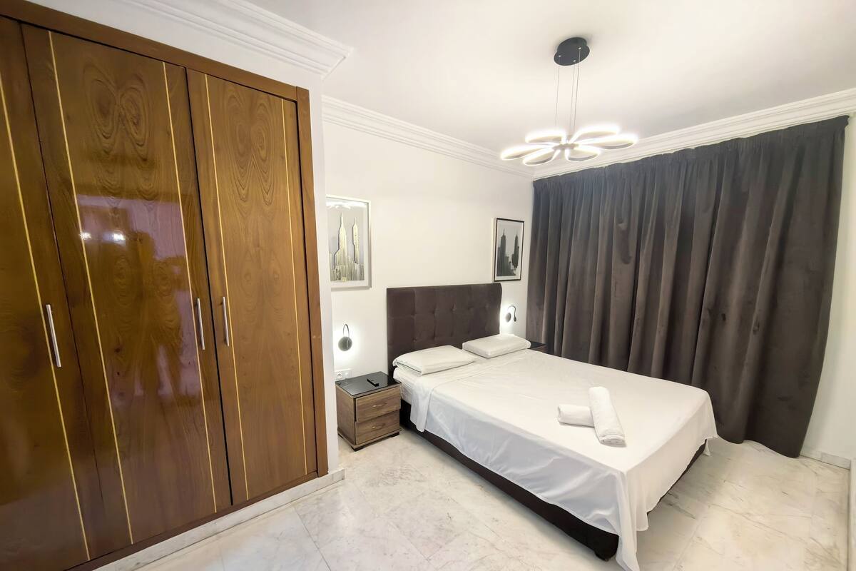 A hotel room with a large wardrobe, a bed with white linens, a nightstand with a lamp, and a modern ceiling light fixture.