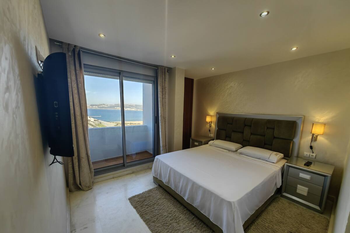 A hotel room with a large bed, a nightstand with a lamp, a TV mounted on the wall, and a view of the sea through a large window.