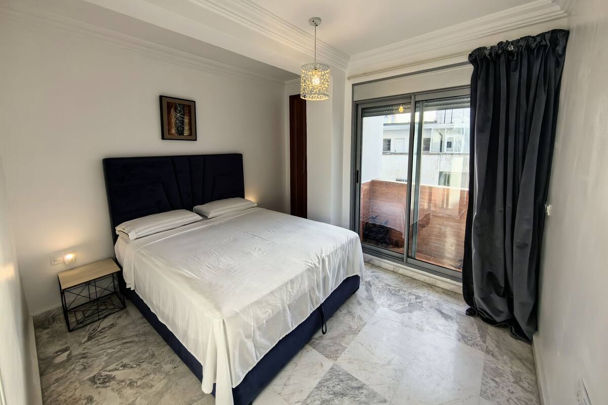 A bedroom with a bed, a bedside table, a hanging light fixture, and a view of the outside.