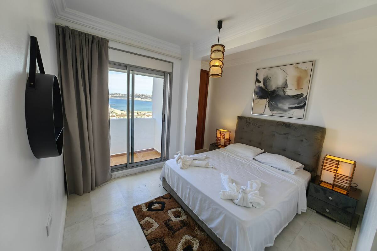 A bedroom with a bed, a TV, a painting, and a view of the sea.