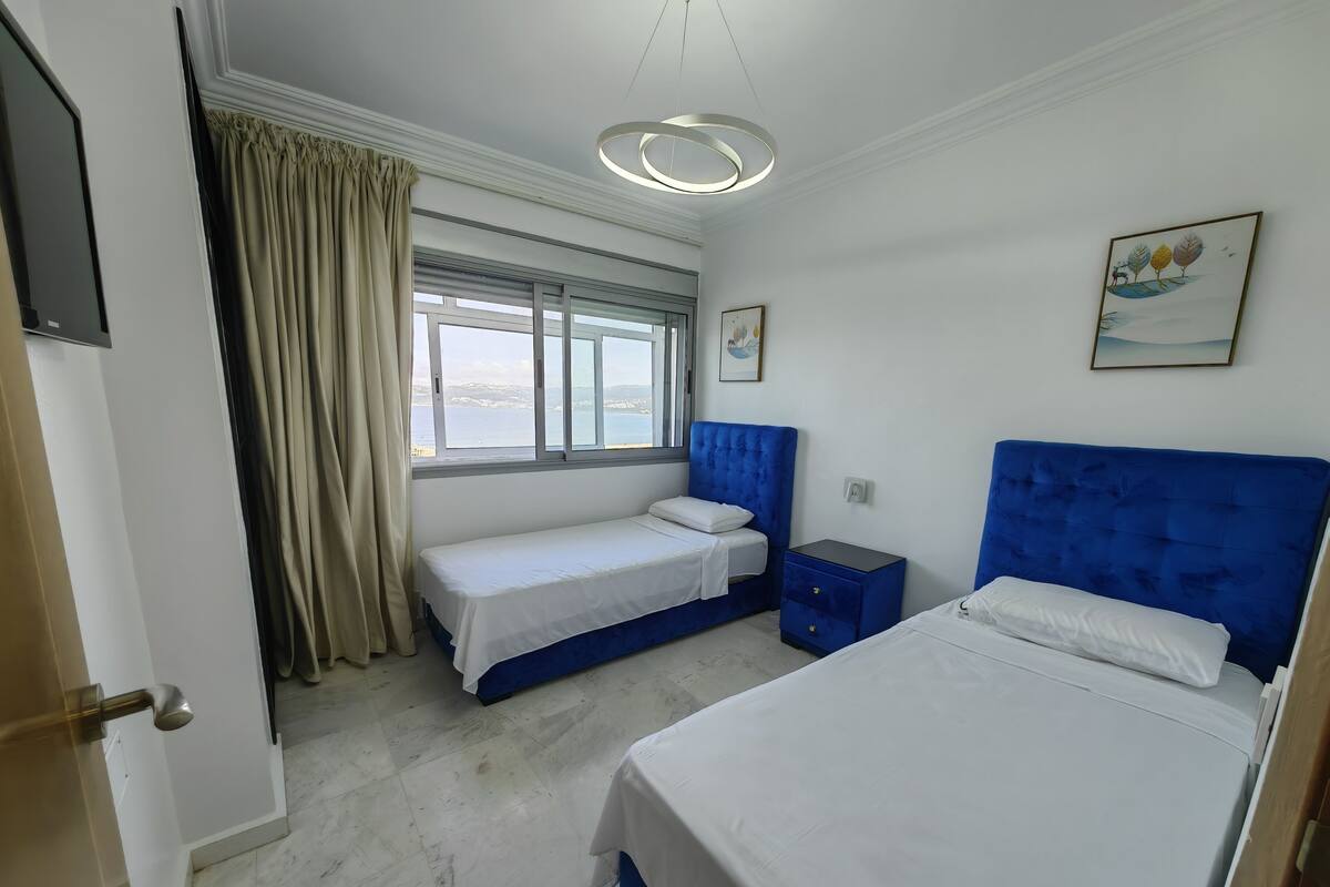 A bedroom with two beds, a window with curtains, a bedside table, and a painting on the wall.