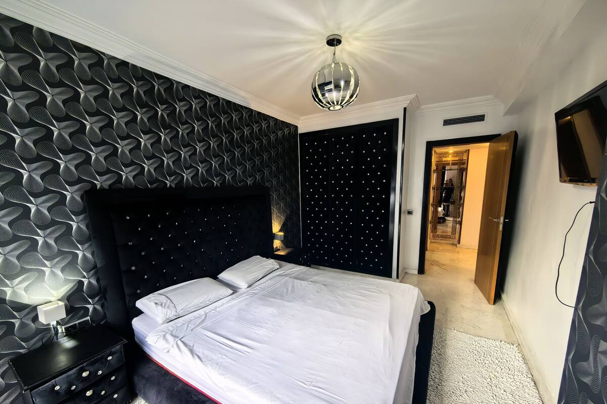 A modern bedroom with a black and white patterned wall, a black headboard with buttons, a white bedspread, and a red accent on the bed.
