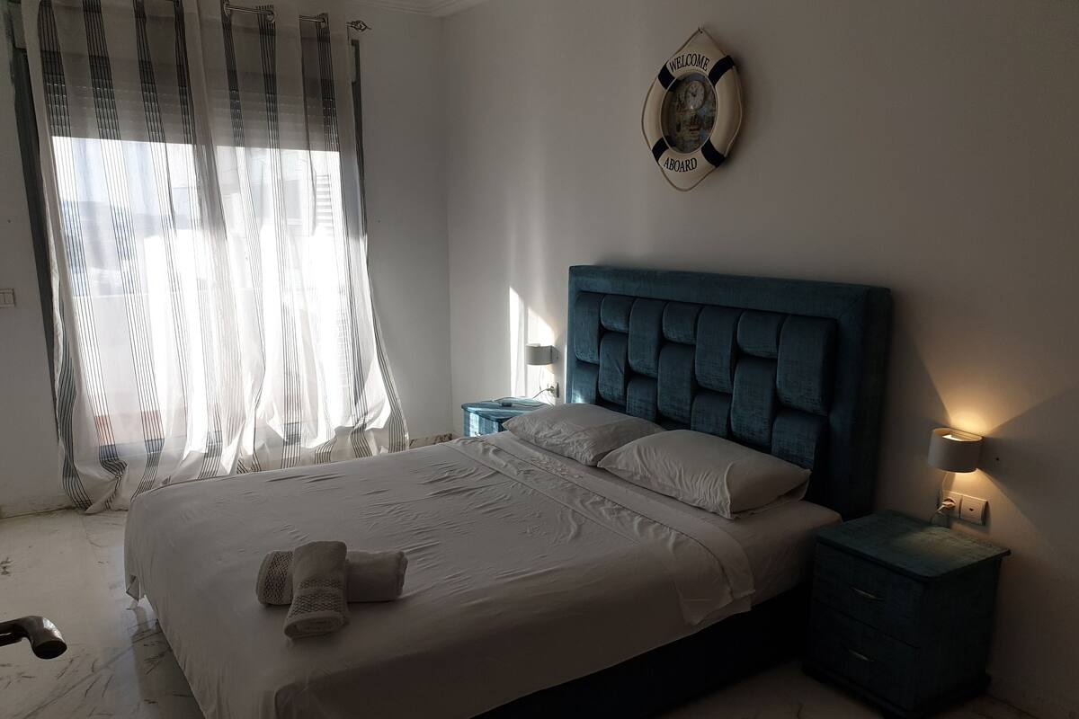 A bedroom with a bed, a nightstand, a wall clock, and sheer curtains.