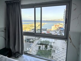 A hotel room with a large window overlooking a coastal area, including a beach and a marina.