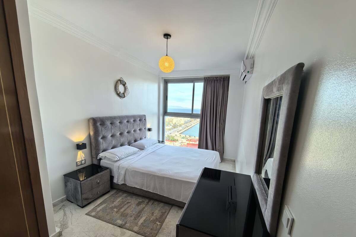 A modern bedroom with a large bed, a bedside table with a lamp, a TV, and a view of the city through a window.