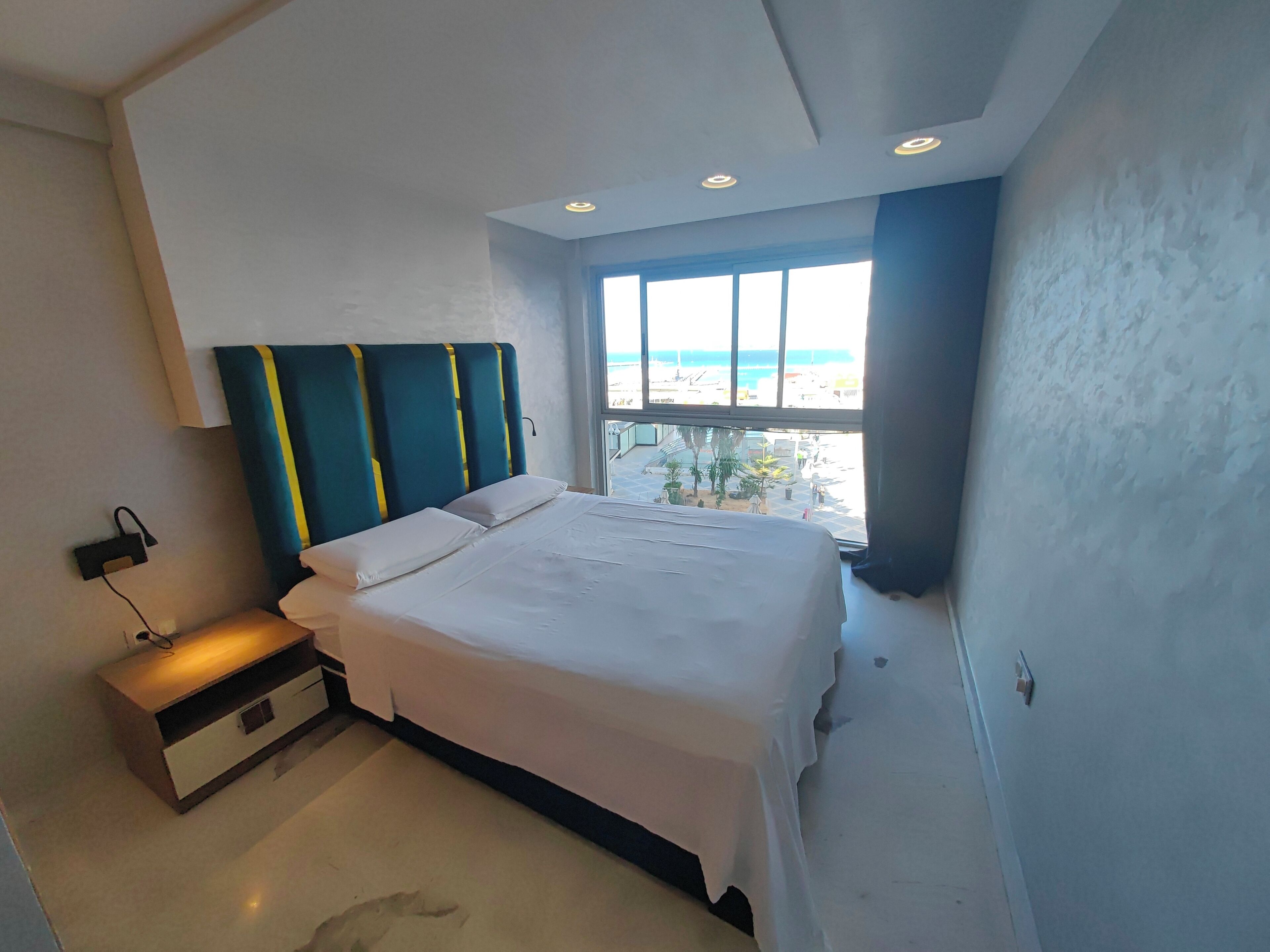 A modern bedroom with a large bed, a nightstand, and a window with a view of the beach.