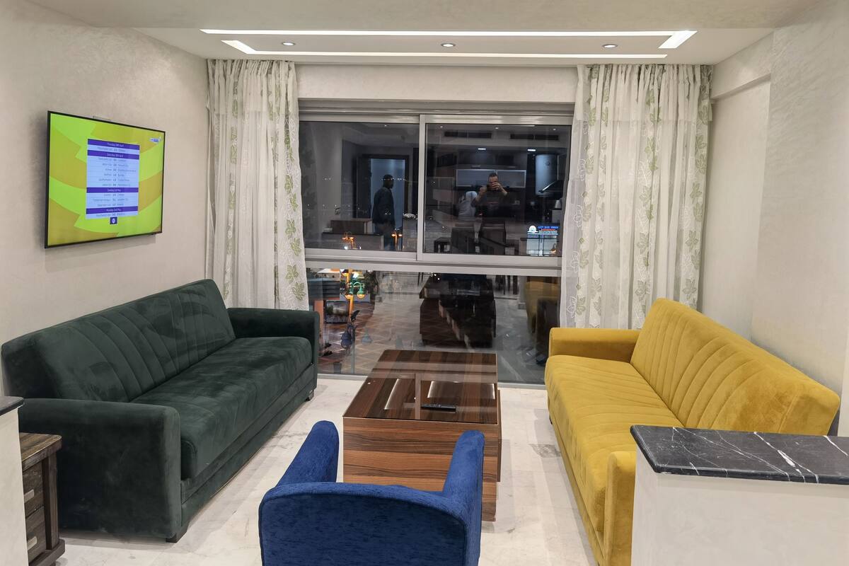 A living room with a green sofa, a yellow sofa, and a blue armchair. There is a TV mounted on the wall and a window with curtains.