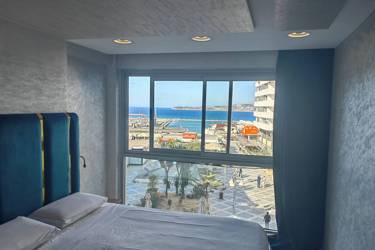 A hotel room with a large window offering a view of the sea and a cityscape.
