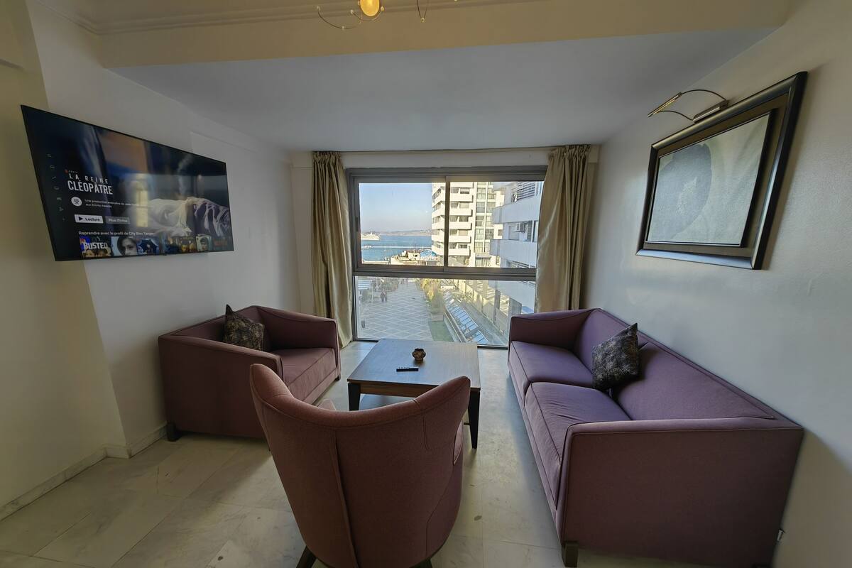 A modern living room with a sofa, armchair, and coffee table. A large window offers a view of a cityscape and a body of water.