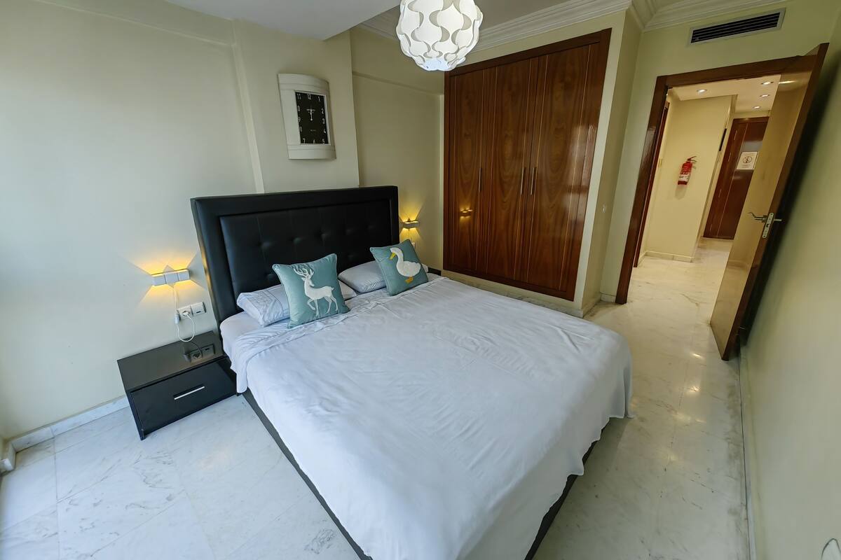A bedroom with a large bed, a wooden wardrobe, a nightstand, and a wall-mounted clock.