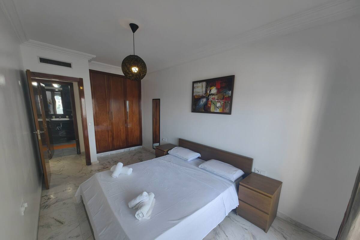 A hotel room with a large bed, wooden wardrobe, marble flooring, and a ceiling light fixture.