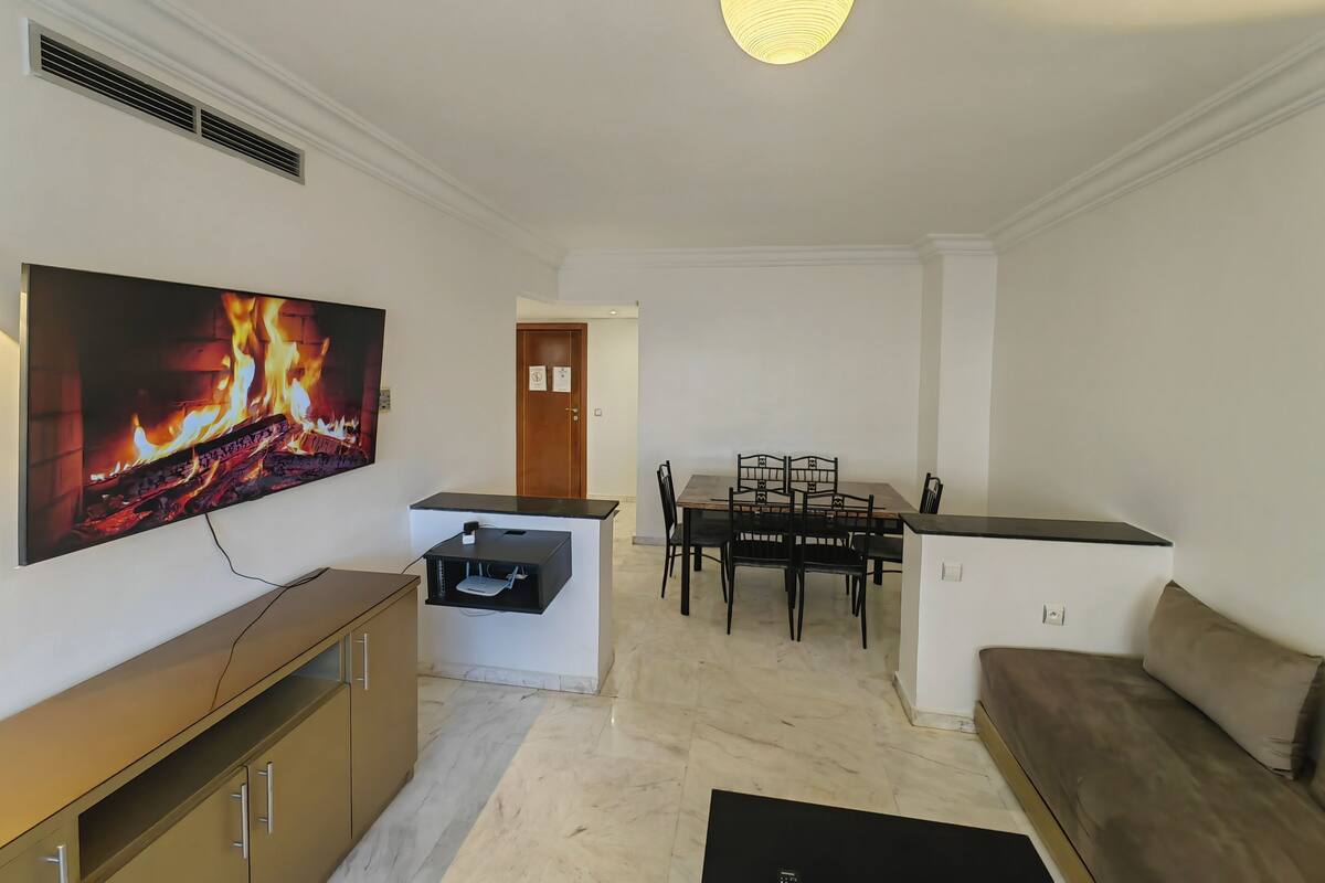 A modern living room with a flat-screen TV, a dining table with chairs, a sofa, and a ceiling light fixture.