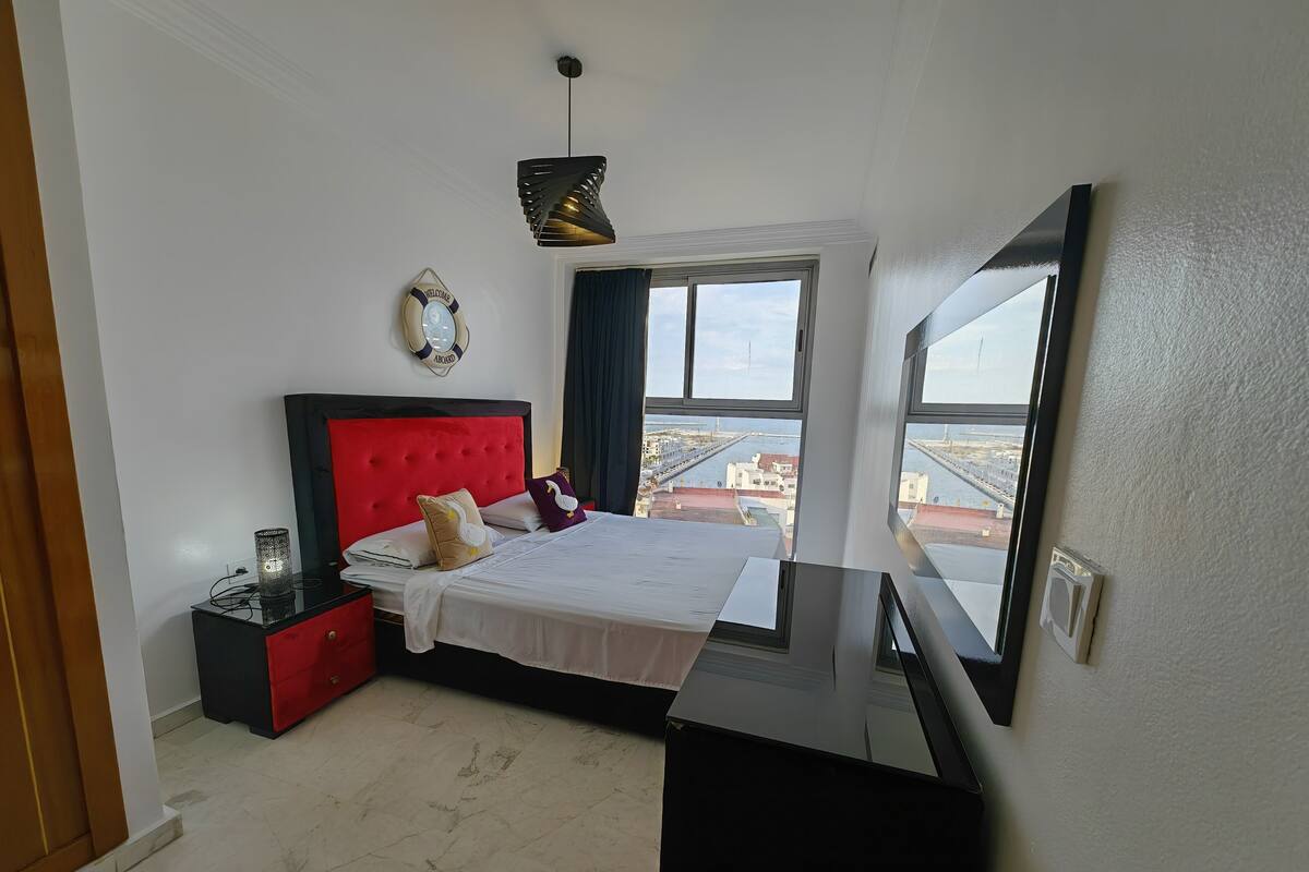 A bedroom with a red headboard, a bed with white linens, a bedside table with a lamp, and a view of the city through large windows.