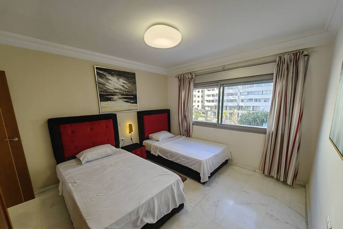 A hotel room with two beds, a painting on the wall, a window with curtains, and a view of buildings outside.