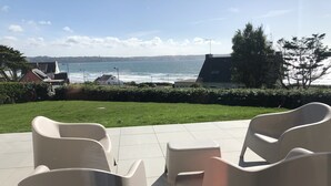 Eine Terrasse mit modernen Stühlen und einem kleinen Tisch, mit Blick auf die Küste, ein Rasenstück und Gebäude im Hintergrund.