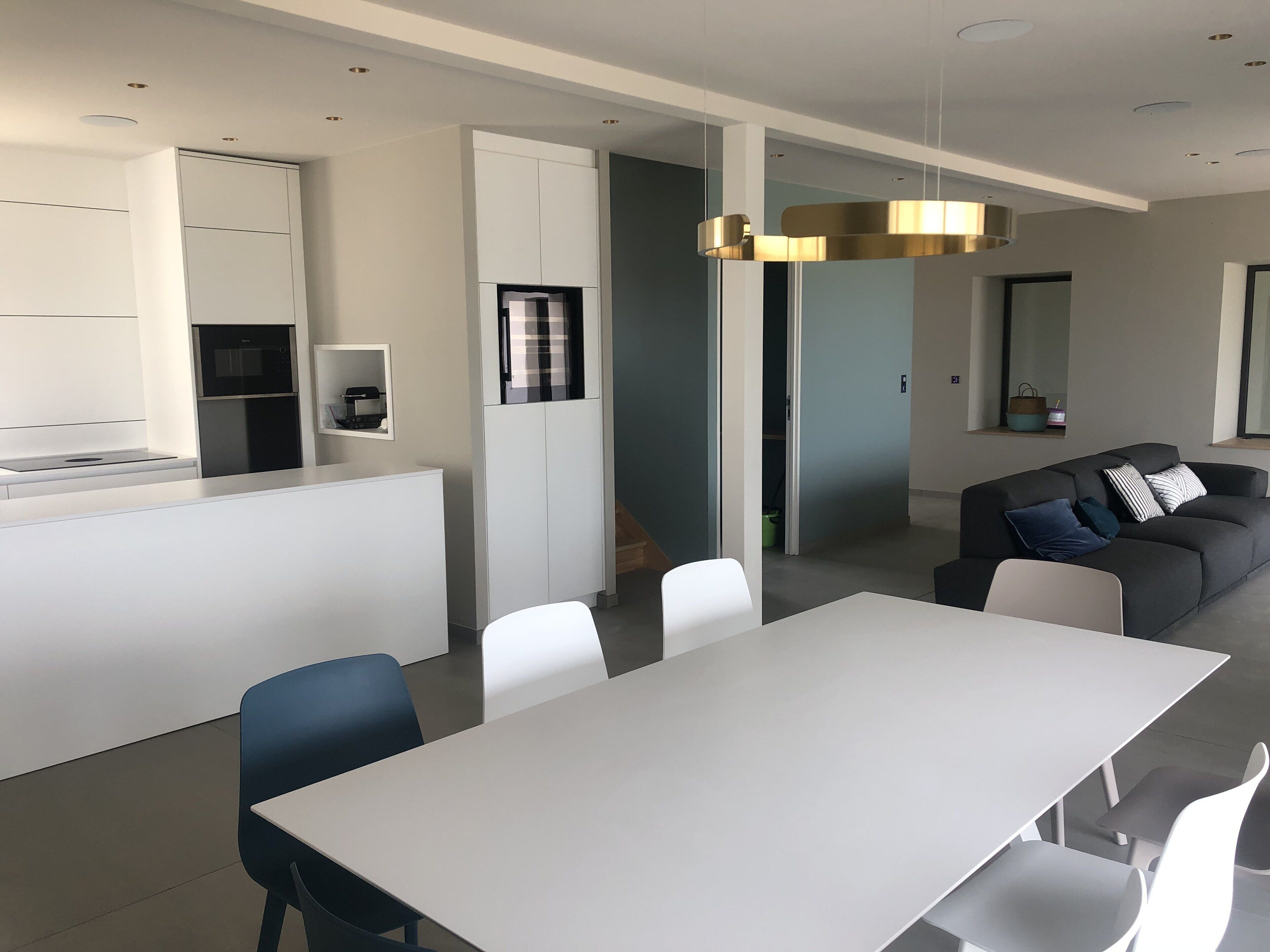 A modern kitchen with white cabinetry, a built-in oven, and a microwave. There is a dining area with a white table and chairs, and a grey sofa with cushions.