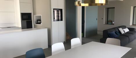 A modern kitchen with white cabinetry, a built-in oven, and a microwave. There is a dining area with a white table and chairs, and a grey sofa with cushions.