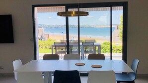 Ein Essbereich mit Tisch und Stühlen, große Fenster mit Blick auf Strand und Meer sowie eine Pendelleuchte über dem Tisch.