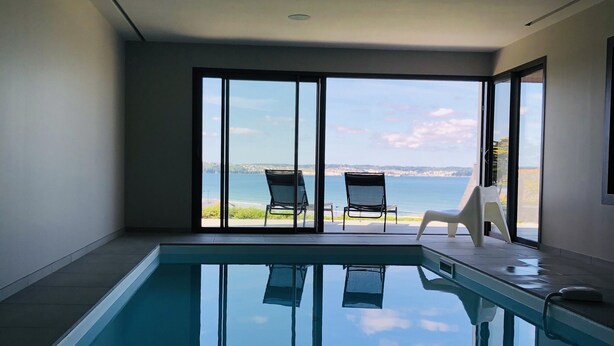 Ein moderner, überdachter Pool mit Panoramafenstern, zwei Liegestühlen und Blick auf das Meer.