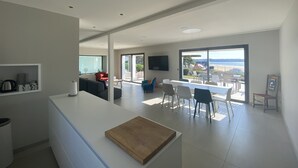 Ein modernes Wohnzimmer mit einer großen Schiebeglastür, die auf einen Balkon führt, einem Essbereich mit weißem Tisch und Stühlen sowie einer Küchenzeile mit weißer Arbeitsplatte und Schneidebrett.