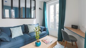 A modern living room with a blue sofa, a coffee table, a potted plant, and a desk with a chair. - Le Matissia (Paris)