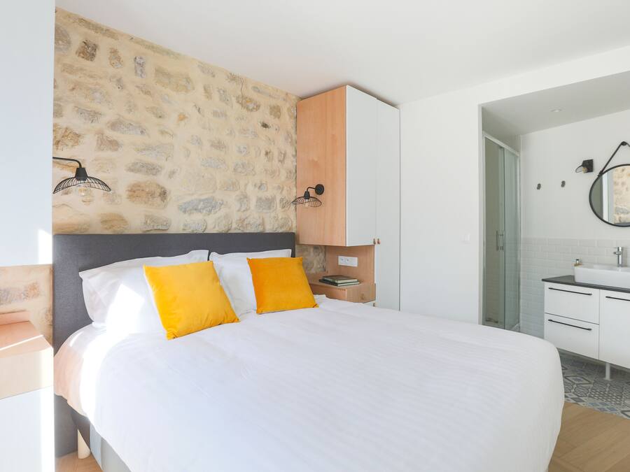 A bedroom with a stone wall, a bed with white bedding and yellow pillows, a wooden cabinet, a bedside table, and a bathroom with a sink and mirror.