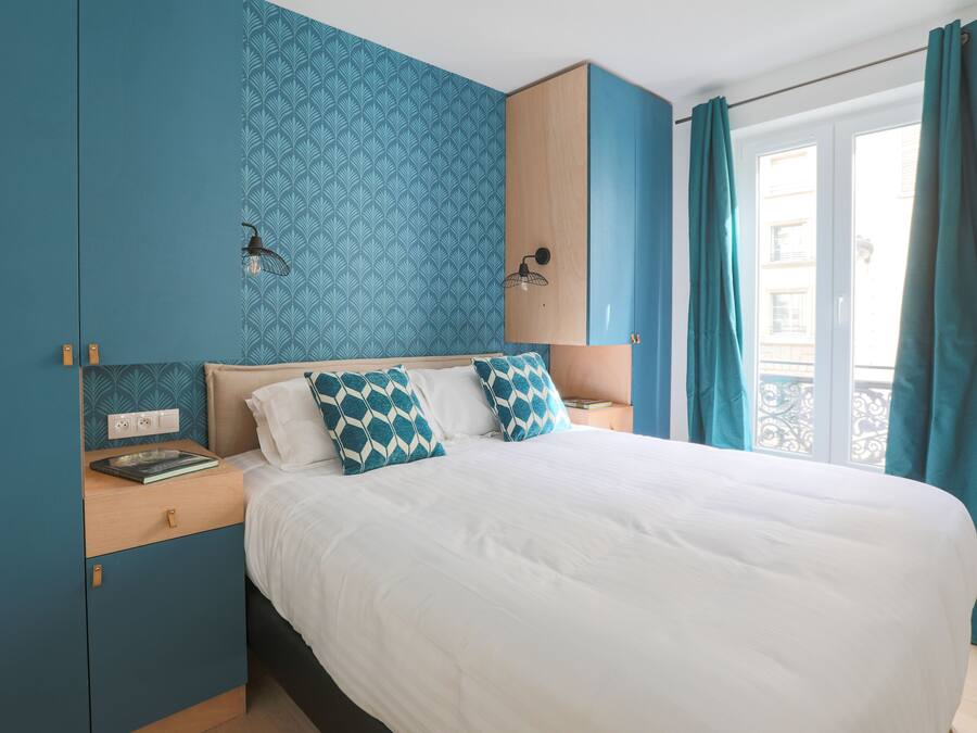 A neatly made bed with white bedding and patterned pillows, a bedside table with a book, and a wall with a geometric pattern.