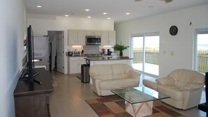 A modern living room with a glass coffee table, two white sofas, a TV, and a kitchen with white cabinets and a microwave. - Golden Surfside--It has GOLDEN VIEWS and it's brand new!! (Surfside Beach)