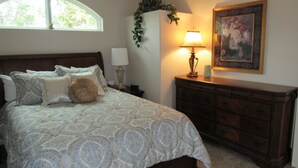 Une chambre à coucher avec un grand lit, une commode, une table de chevet, une lampe et une fenêtre donnant sur des arbres.