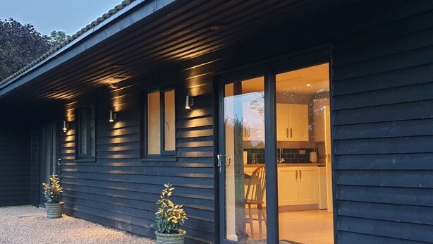 A modern house with a dark exterior, large sliding glass doors leading to a kitchen area with cabinets and a chair visible. - Swallows Nest, Cottage, Fishing Lakes Nr Cleethorpes, Adults Only Pet Friendly  (Lincolnshire)