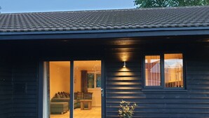 A dark wooden building with a tiled roof, a sliding glass door leading to an interior with a sofa and a small table, and a potted plant outside. - Swallows Nest, Cottage, Fishing Lakes Nr Cleethorpes, Adults Only Pet Friendly  (Lincolnshire)