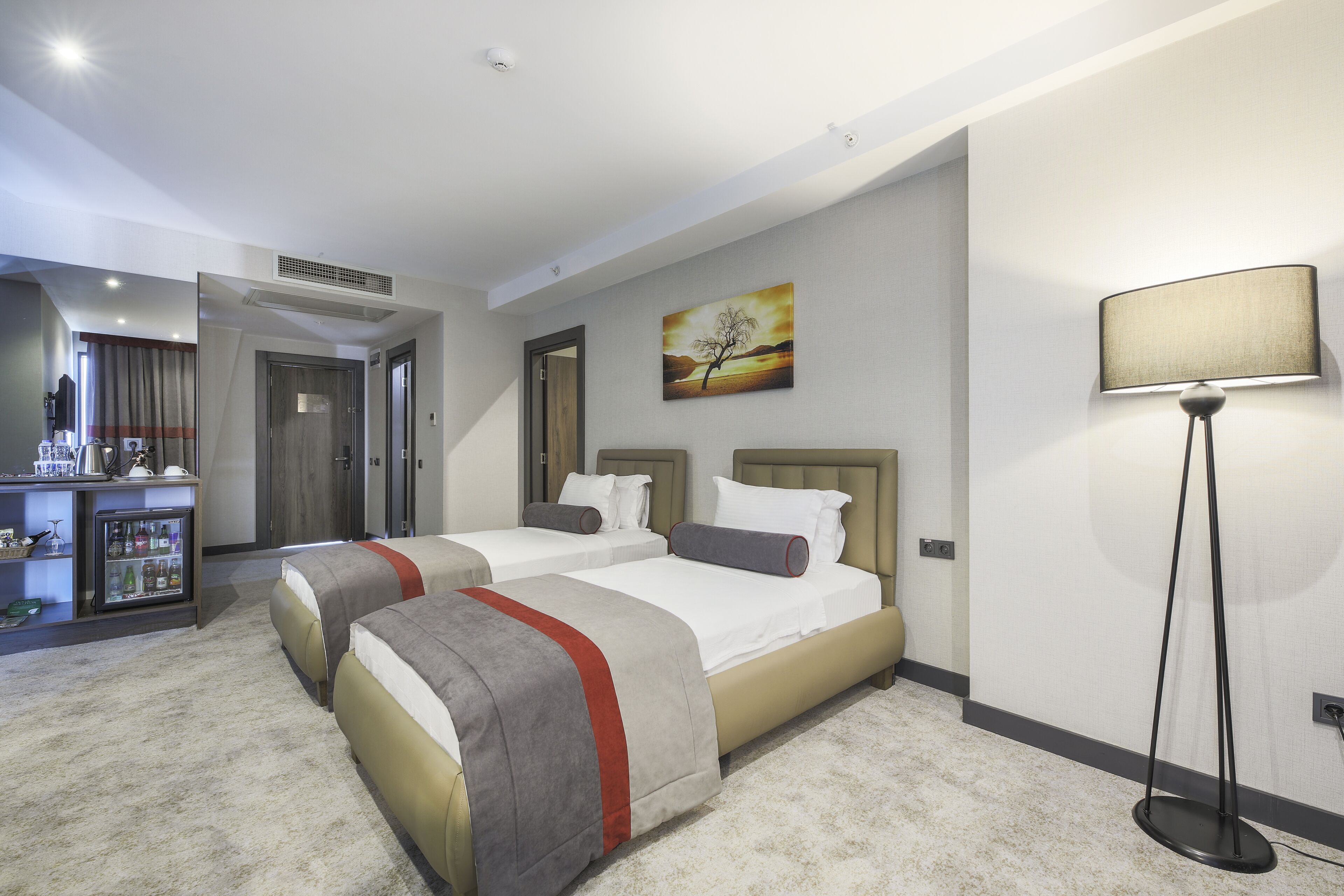 A hotel room with two beds, a desk with a refrigerator, and a painting on the wall.