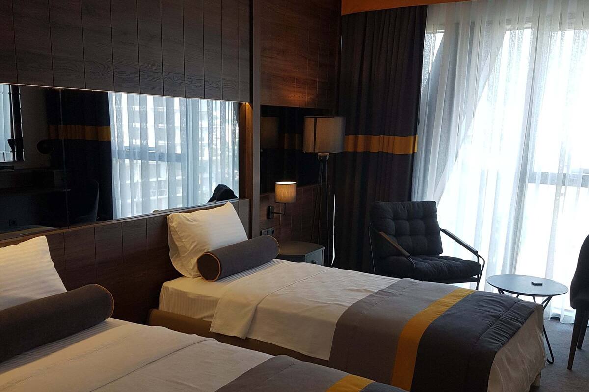 A hotel room with two beds, a desk, a chair, a TV, and a large window with curtains.