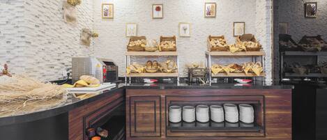 A bakery counter with bread and pastries, a coffee machine, and a display of plates and cups.