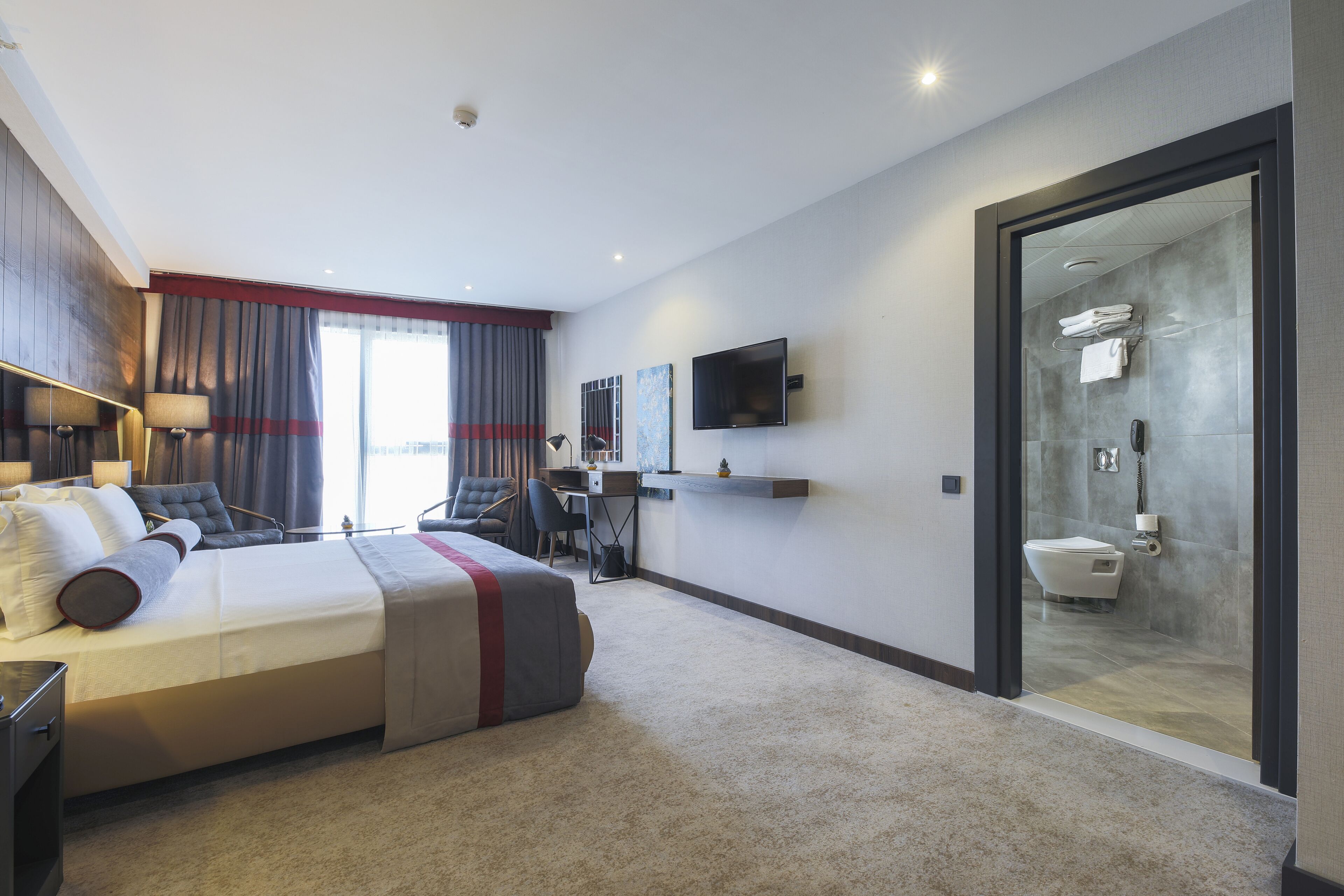 Una habitación de hotel moderna con una cama grande, un escritorio, una silla, un televisor y zona de ducha con paredes de vidrio.