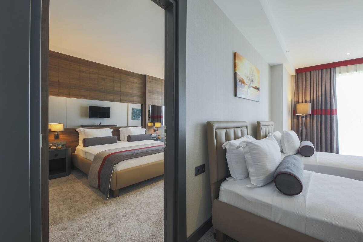 A hotel room with two beds, a flat-screen TV, a painting, and a window with curtains.