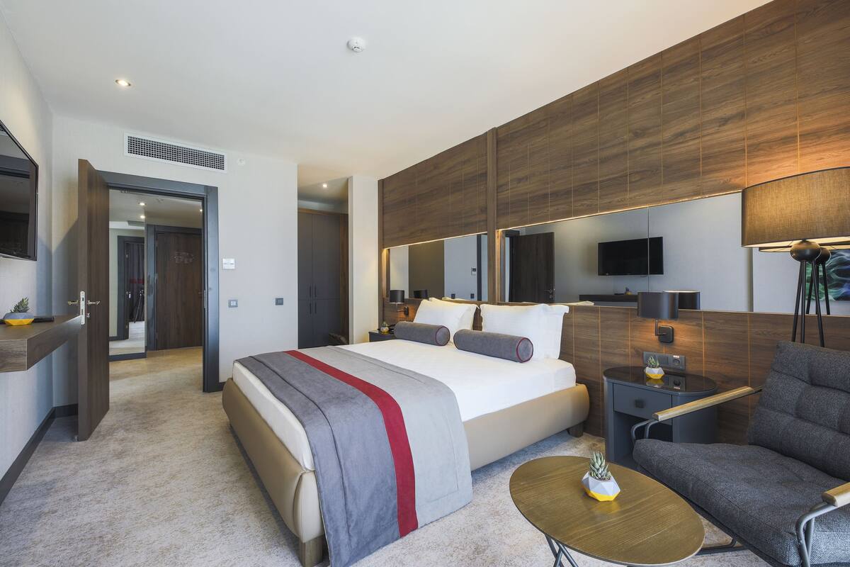 A modern hotel room with a large bed, a sofa, a TV, and a desk with a lamp.