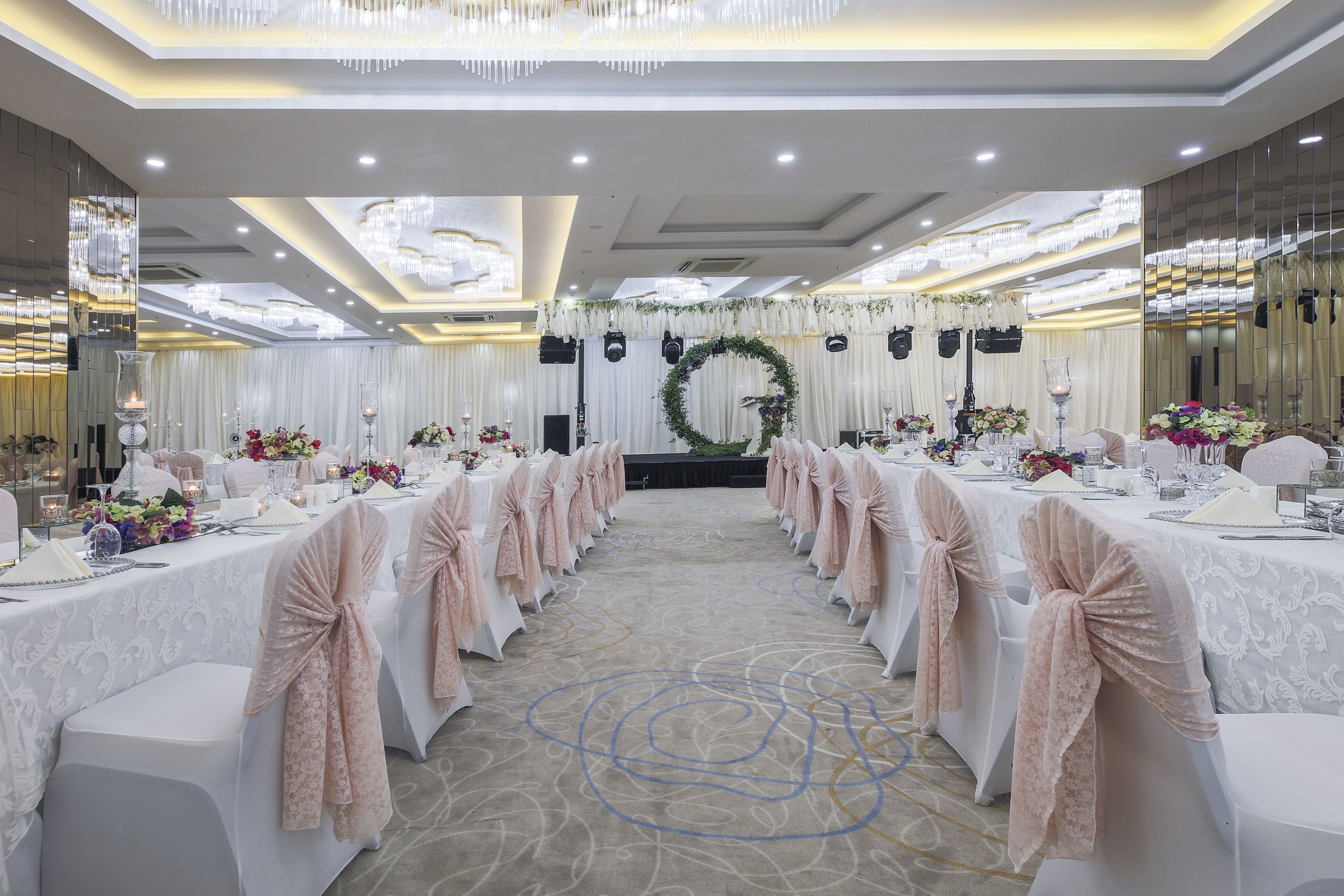 A banquet hall with tables set for a formal event, featuring a central stage, floral decorations, and a chandelier.